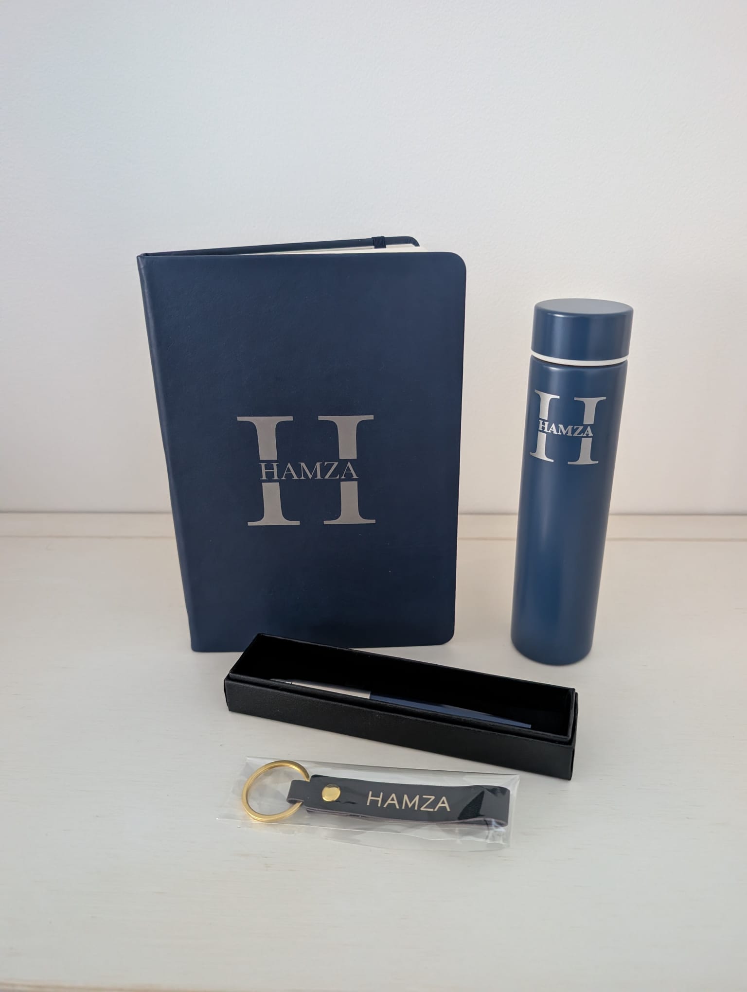 Navy hardback notebook, navy insulated flask, pen in an open black presentation box and navy leather keyring with a gold ring standing together on a pale wooden surface, each carrying the same silver H monogram with the name HAMZA across it